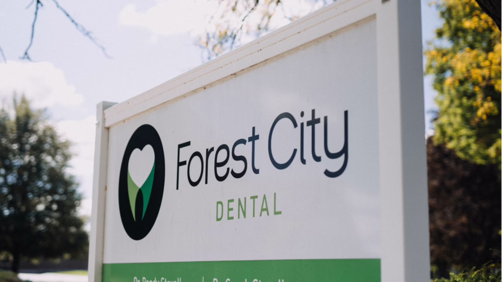 Outdoor sign for Forest City Dental, with a circular logo and green accents, trees in the background.