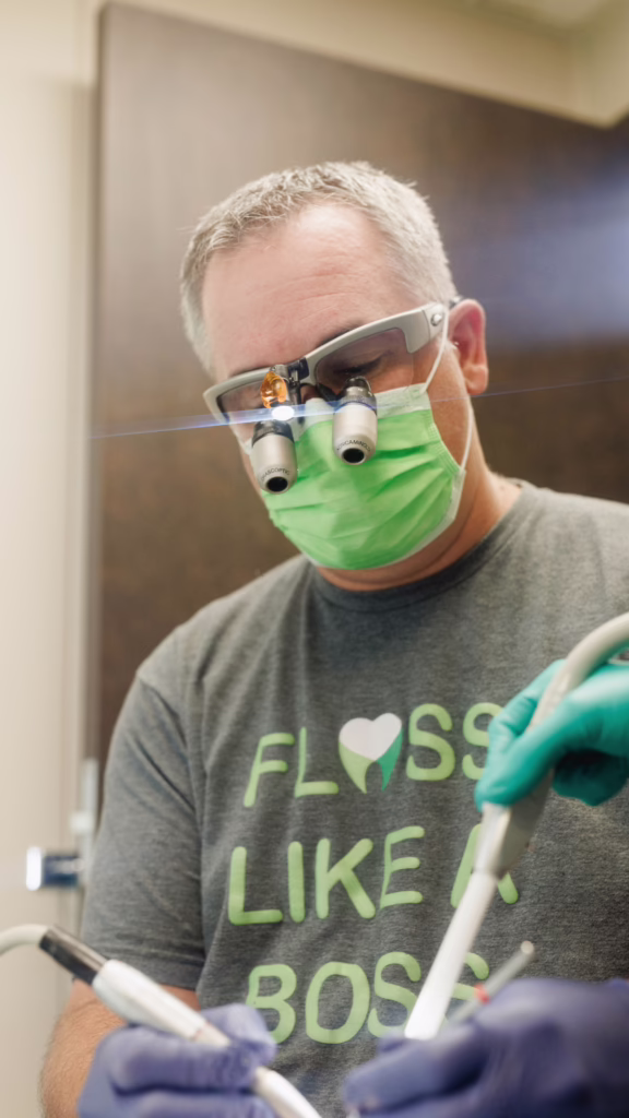 Dental clinician wearing a green mask and head-mounted magnifying loupes, performing a procedure.
