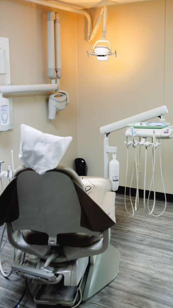 Dental exam chair in a dental clinic with an overhead lamp and instrument tray nearby.”, “Dental office setup: patient chair, dental unit with handpieces, and surrounding equipment.”, “Dental treatment room featuring a reclining chair, tray of tools, and mounted light.”, “Clinic dental chair with attached equipment and a tray holding instruments.”, “Dental operator’s chair with wall-mounted lights and cords for instruments.”