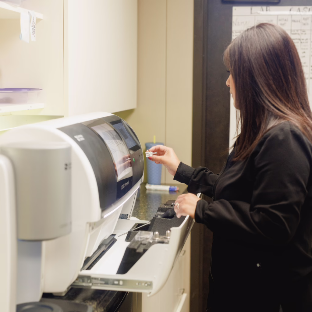 Woman in black shirt inserts a card into a large medical/office device in a clinical setting with cabinets nearby.