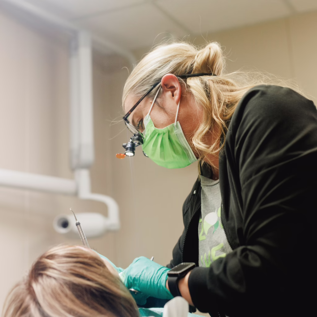 Dentist wearing a green mask and magnifying loupes leans over a patient, using dental tools during a dental procedure in a clinic.