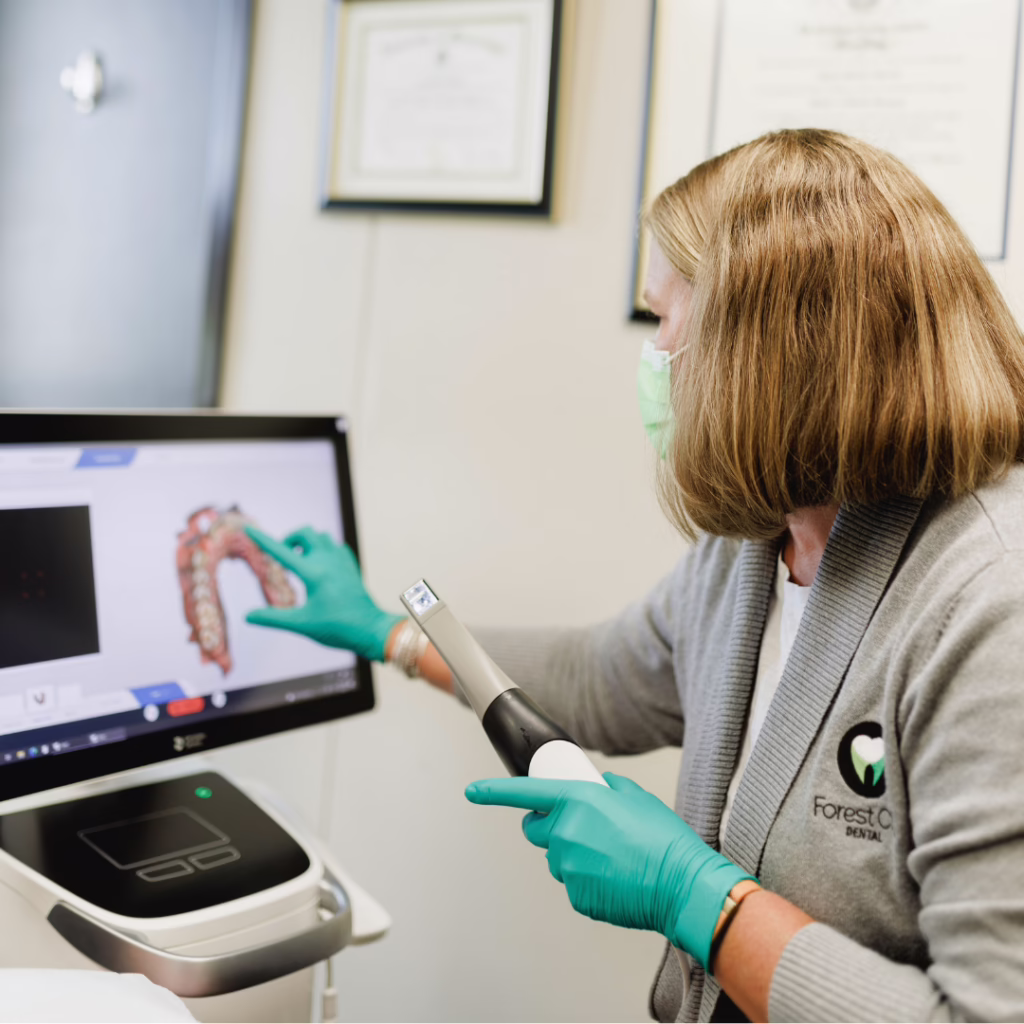 Dental professional in teal gloves and mask uses a handheld intraoral scanner while a 3D dental image appears on the monitor.